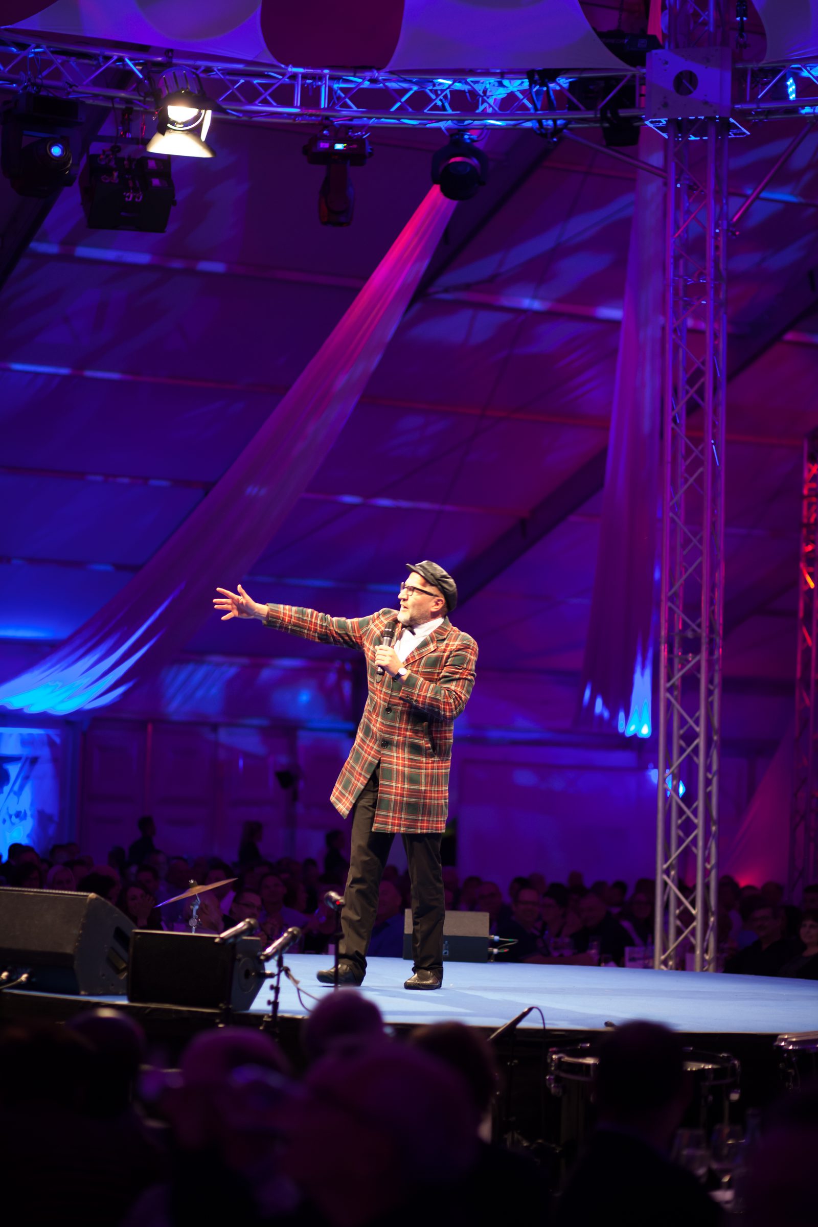 Entertainer in kariertem Mantel und Hut spricht mit Mikrofon auf der violett beleuchteten Bühne im Festzelt beim Firmenjubiläum von Weber Hydraulik