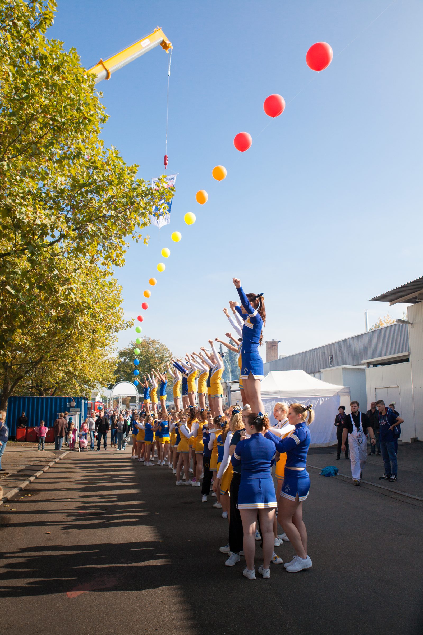Cheerleader-Gruppe zeigt beim Firmenjubiläum von Weber Hydraulik eine Hebefigur auf dem Werksgelände, darüber eine bunte Ballonkette an einem Kran