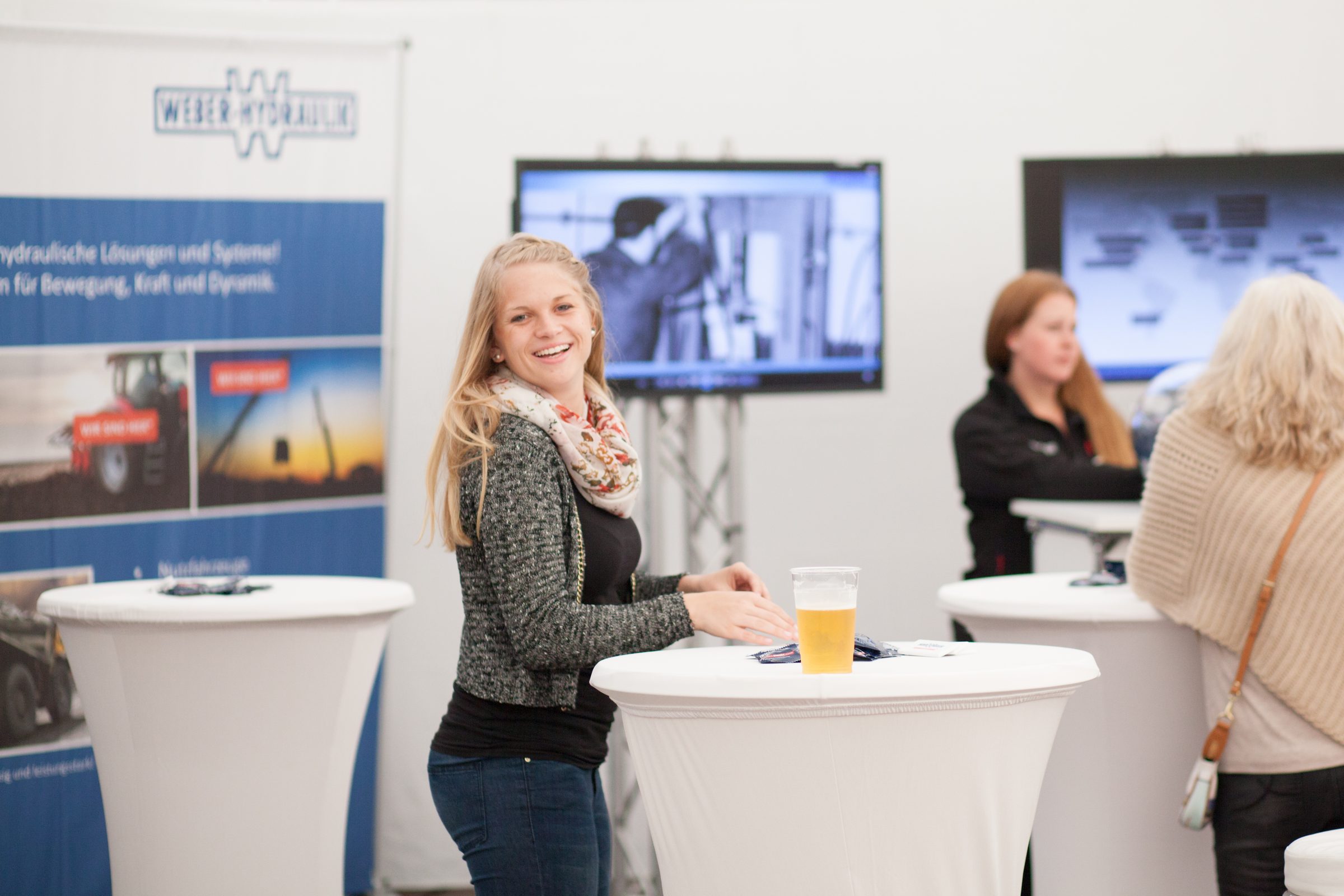 Lächelnde Frau an einem Stehtisch am Stand von Weber Hydraulik, im Hintergrund Bildschirme