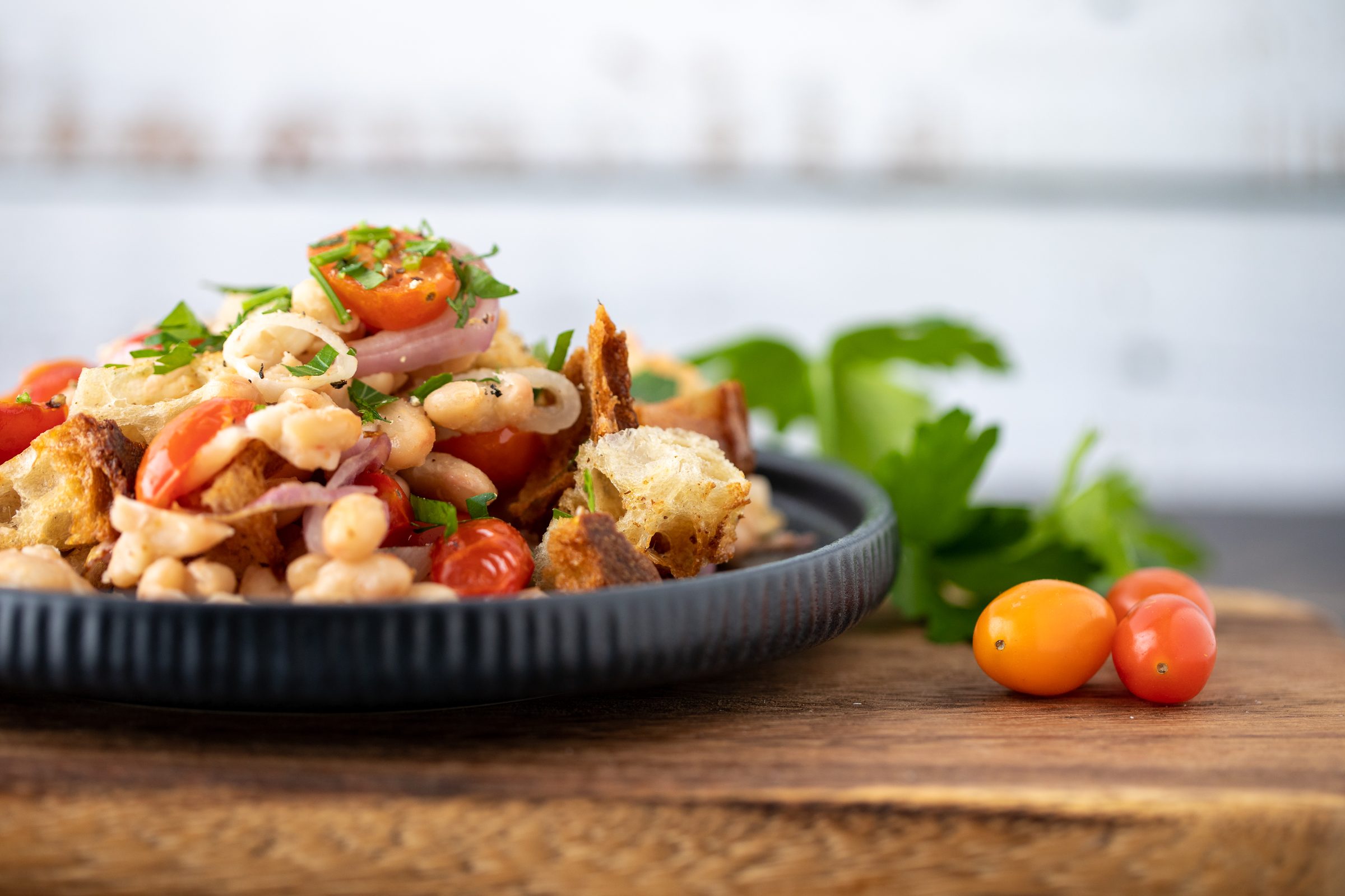 Salat aus weißen Bohnen, gerösteten Brotwürfeln, Tomaten und roten Zwiebeln auf einem dunklen Teller, Rezeptfoto für XbyX