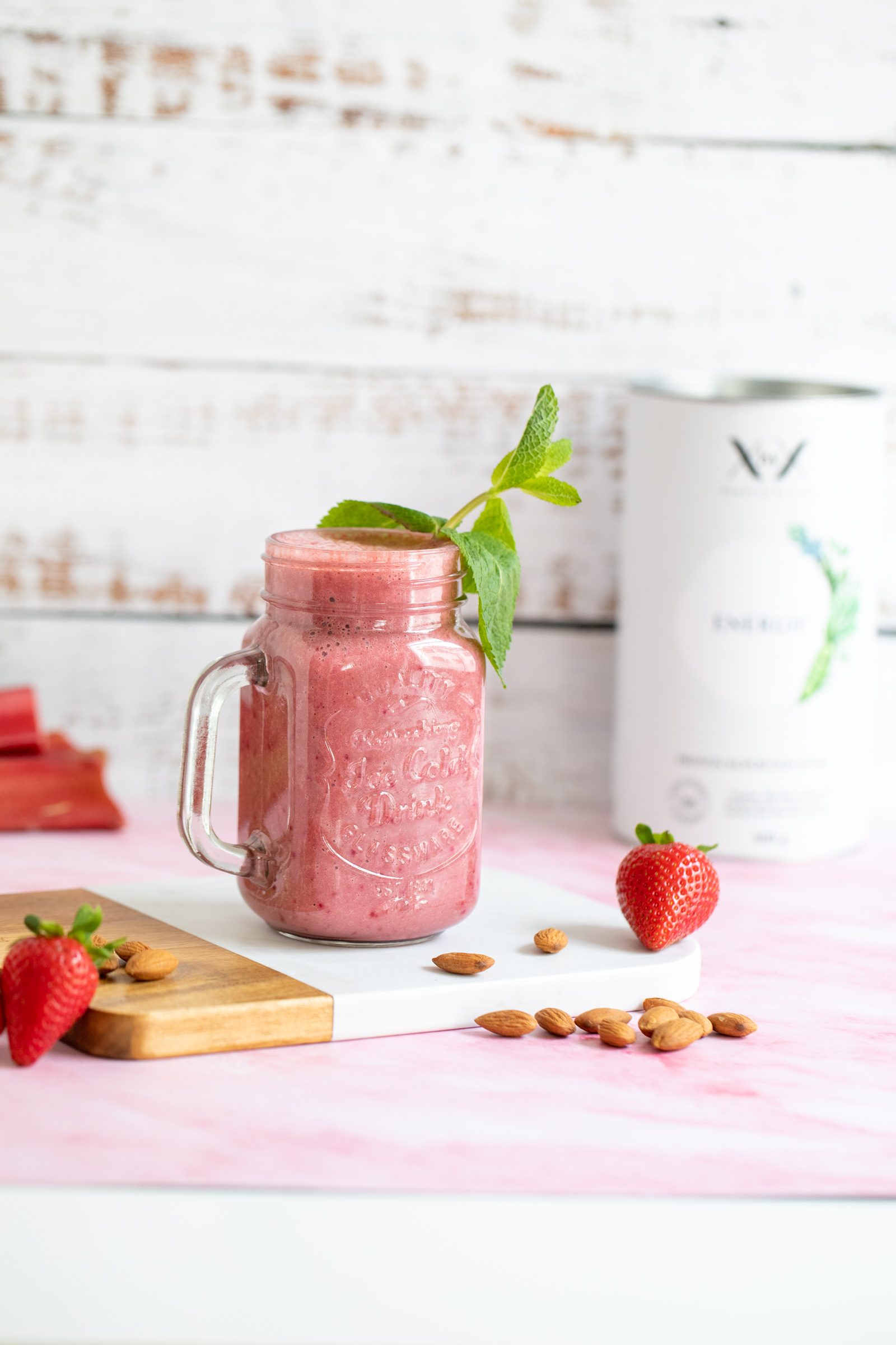 Erdbeer-Rhabarber-Smoothie im Henkelglas mit Minze, daneben Erdbeeren, Mandeln und eine Produktdose von XbyX