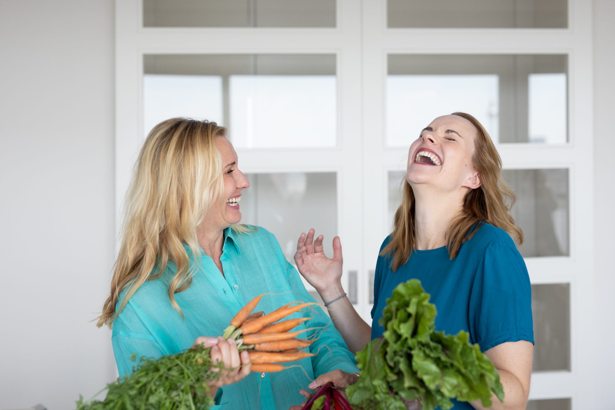 Zwei Frauen lachen gemeinsam vor einer weißen Sprossentür, in den Händen frische Karotten und Mangold, Gründerinnen-Shooting für XbyX