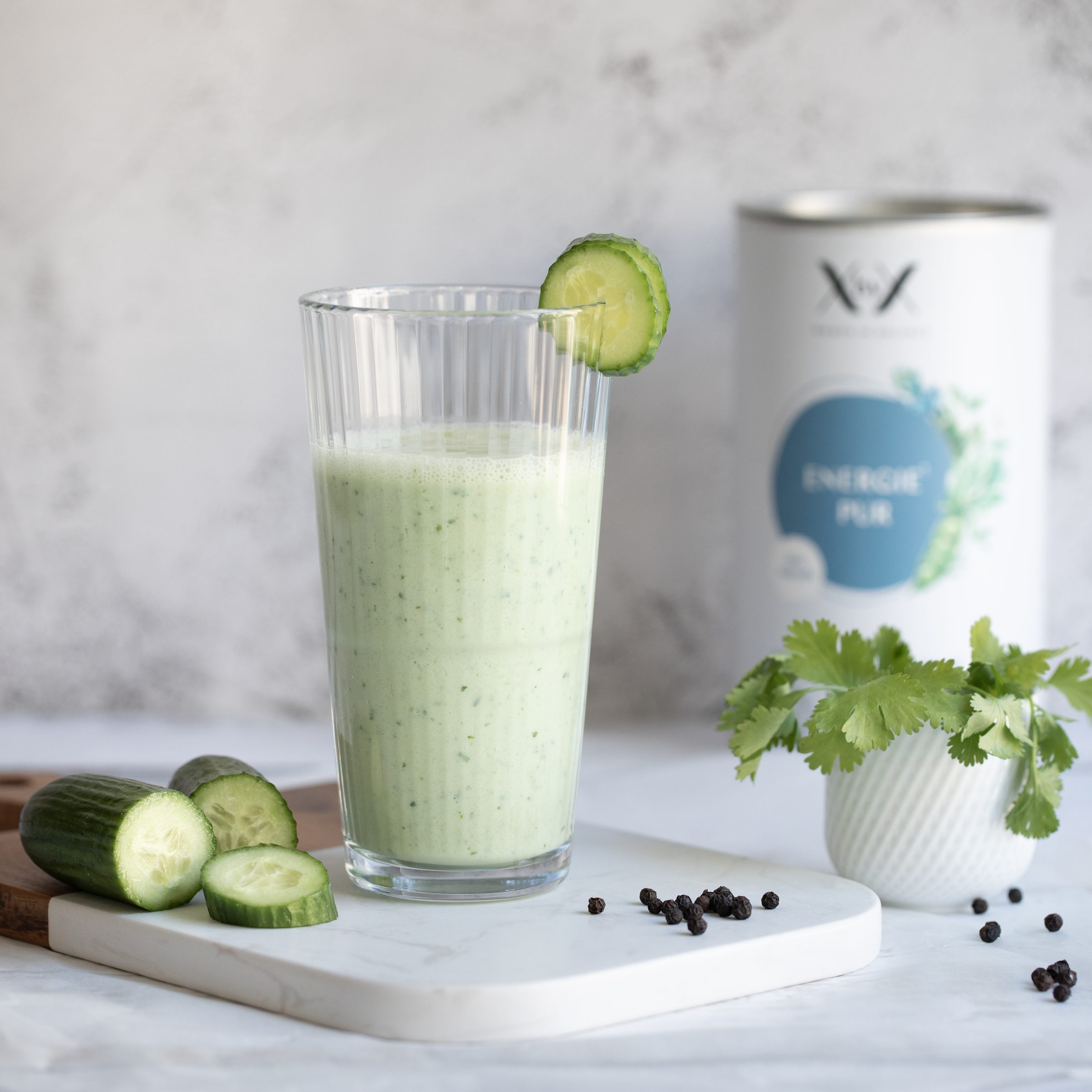 Grüner Gurkensmoothie im hohen Glas mit Gurkenscheibe am Rand, daneben Gurken, Koriander und eine Dose Energie Pur von XbyX