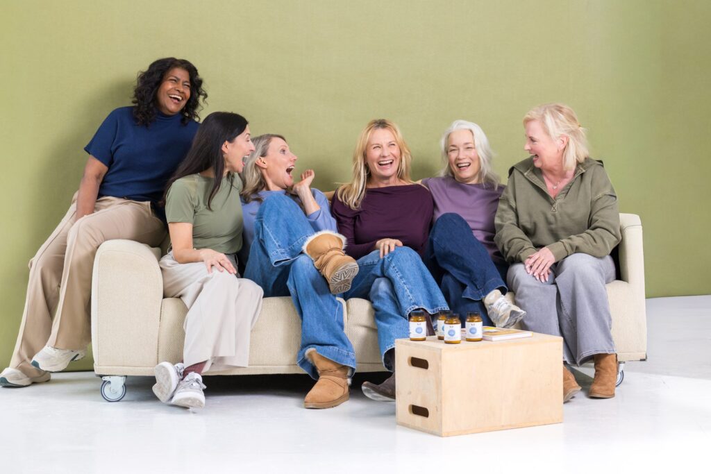 Sechs Frauen sitzen lachend auf einem Sofa vor einer grünen Wand, davor stehen Produkte von XbyX auf einer Holzkiste