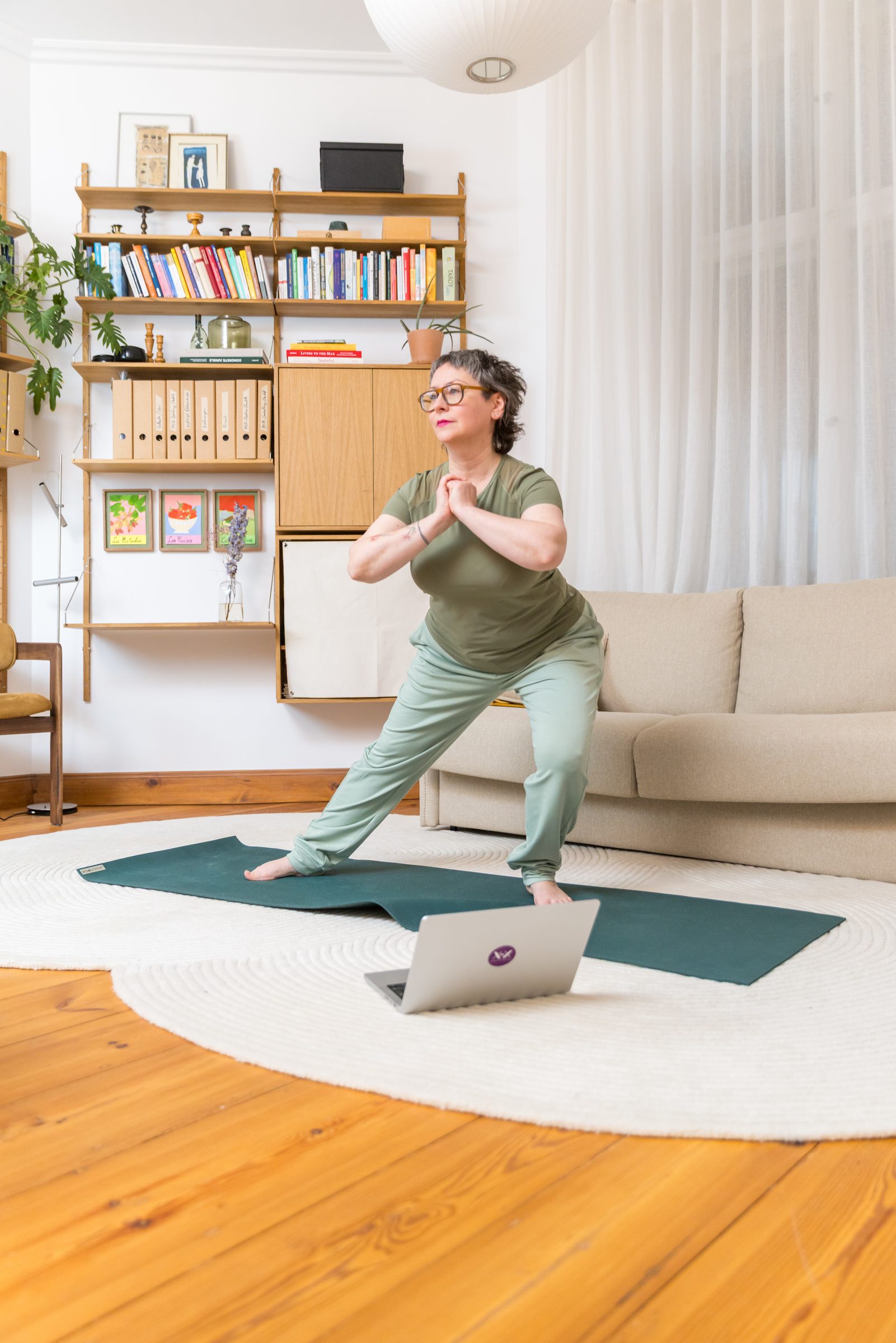 Frau in grüner Sportkleidung macht eine Yogaübung auf einer Matte im Wohnzimmer, vor ihr ein aufgeklappter Laptop, Markenshooting für XbyX