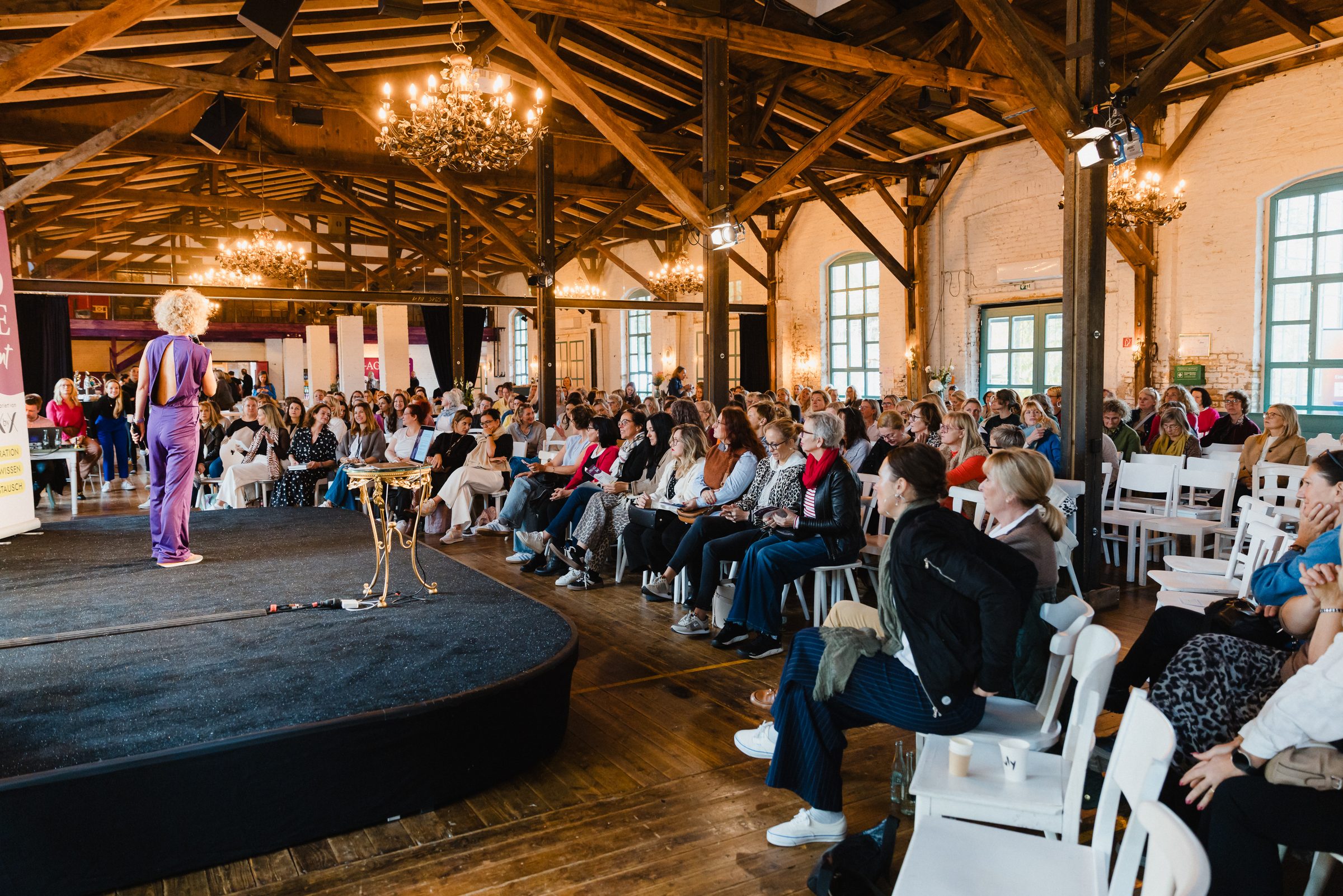Referentin auf der Bühne des Pro Age Event von XbyX vor vollem Saal mit Holzbalken und Kronleuchtern