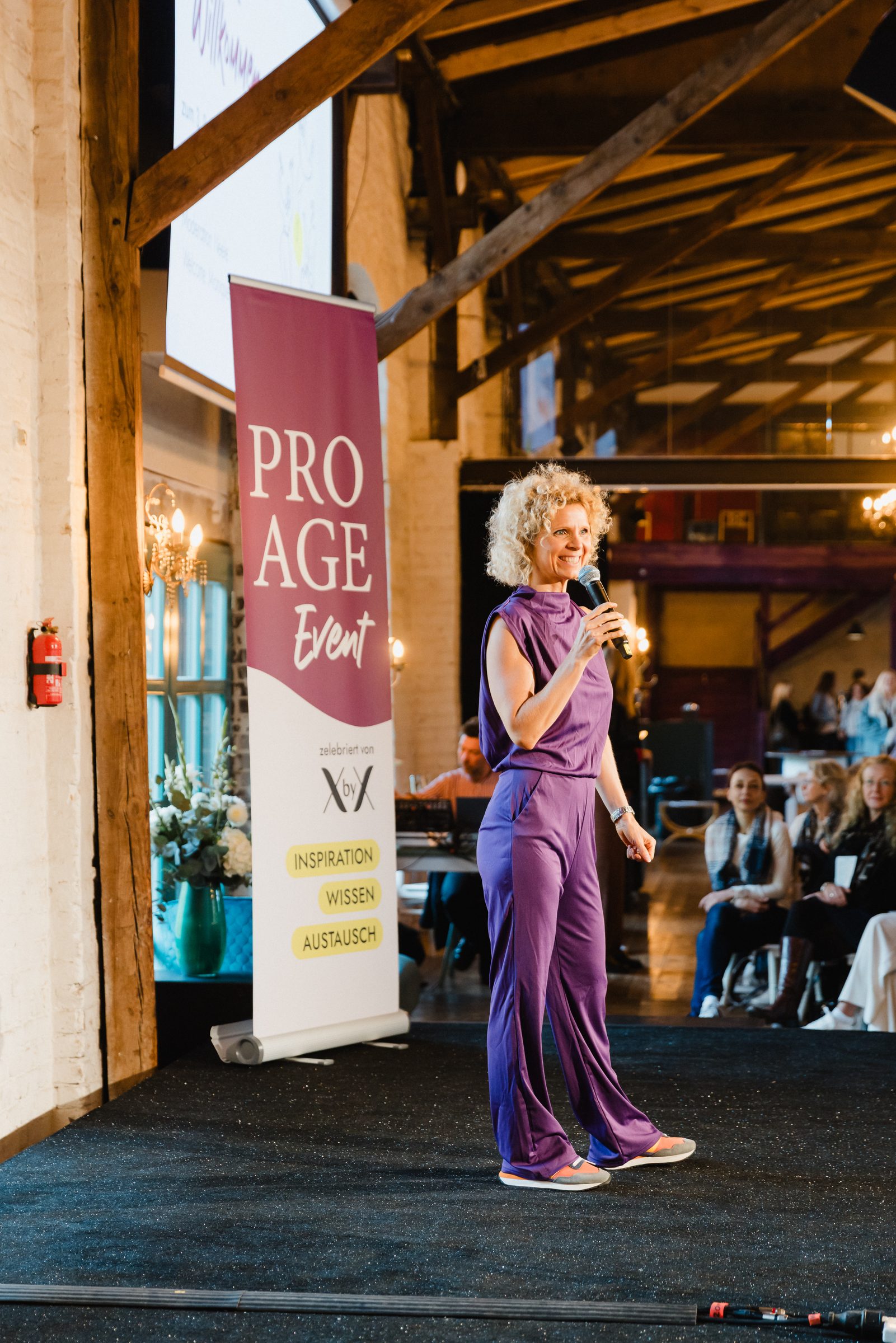 Referentin in lila Anzug spricht mit Mikrofon auf der Bühne beim Pro Age Event von XbyX, daneben ein Roll-up