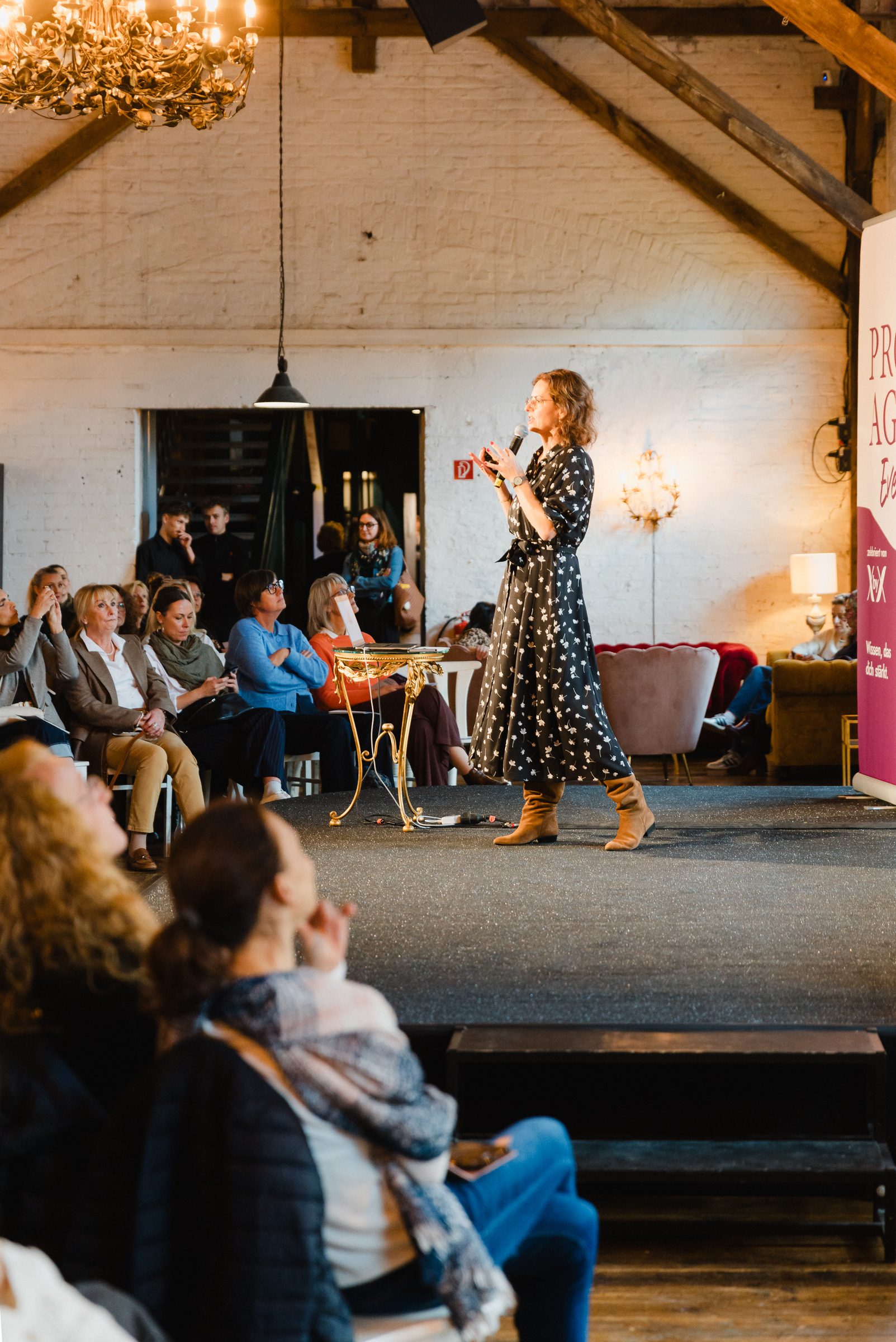 Referentin im gepunkteten Kleid spricht unter einem Kronleuchter vor dem Publikum beim Pro Age Event von XbyX
