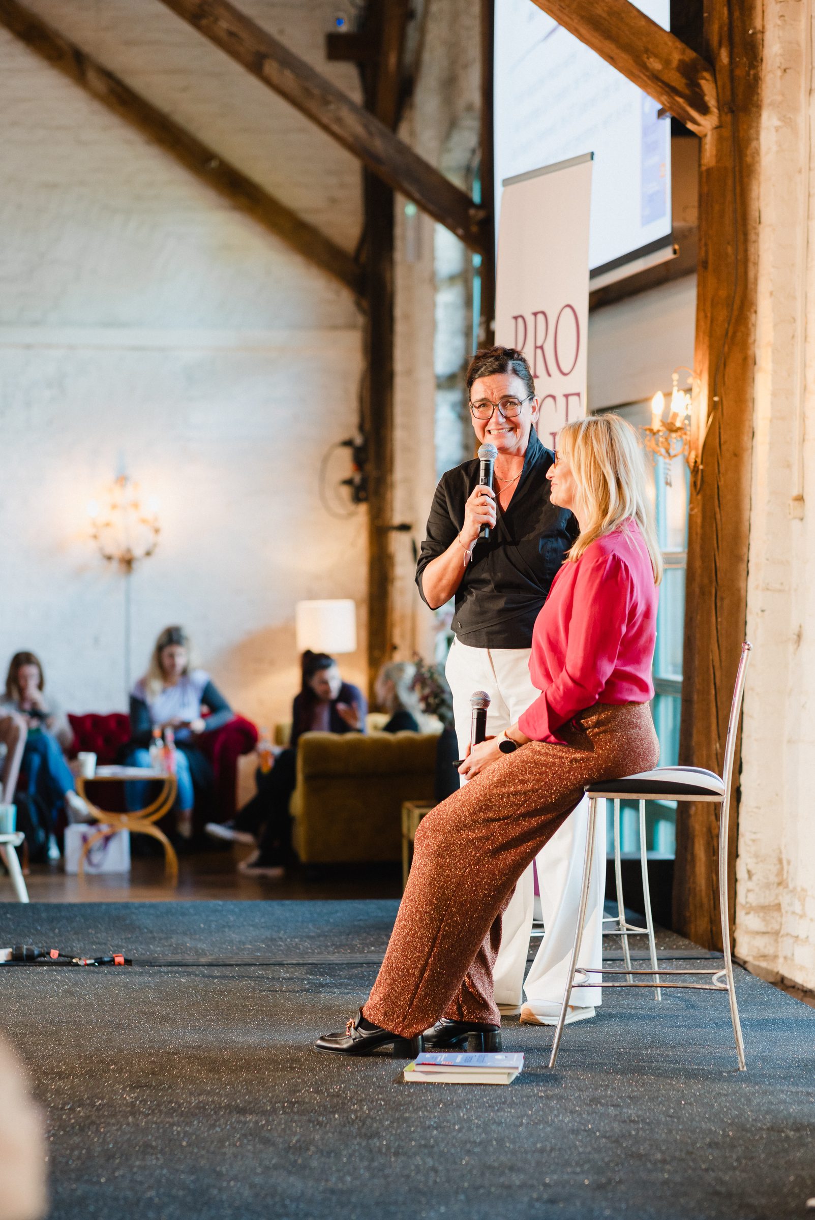 Zwei Frauen im Gespräch auf der Bühne beim Pro Age Event von XbyX, eine spricht ins Mikrofon, die andere sitzt auf einem Barhocker