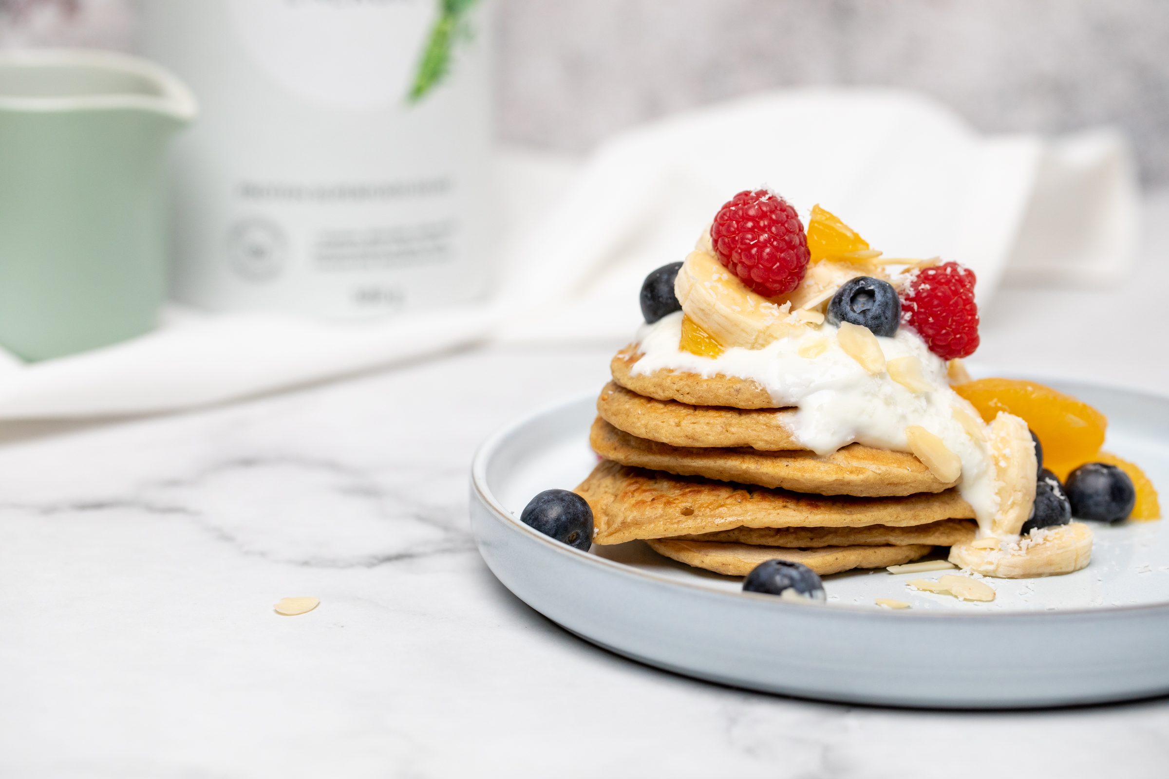 Stapel Protein-Pancakes mit Joghurt, Himbeeren, Heidelbeeren und Bananenscheiben auf einem hellen Teller, Rezeptfoto für XbyX