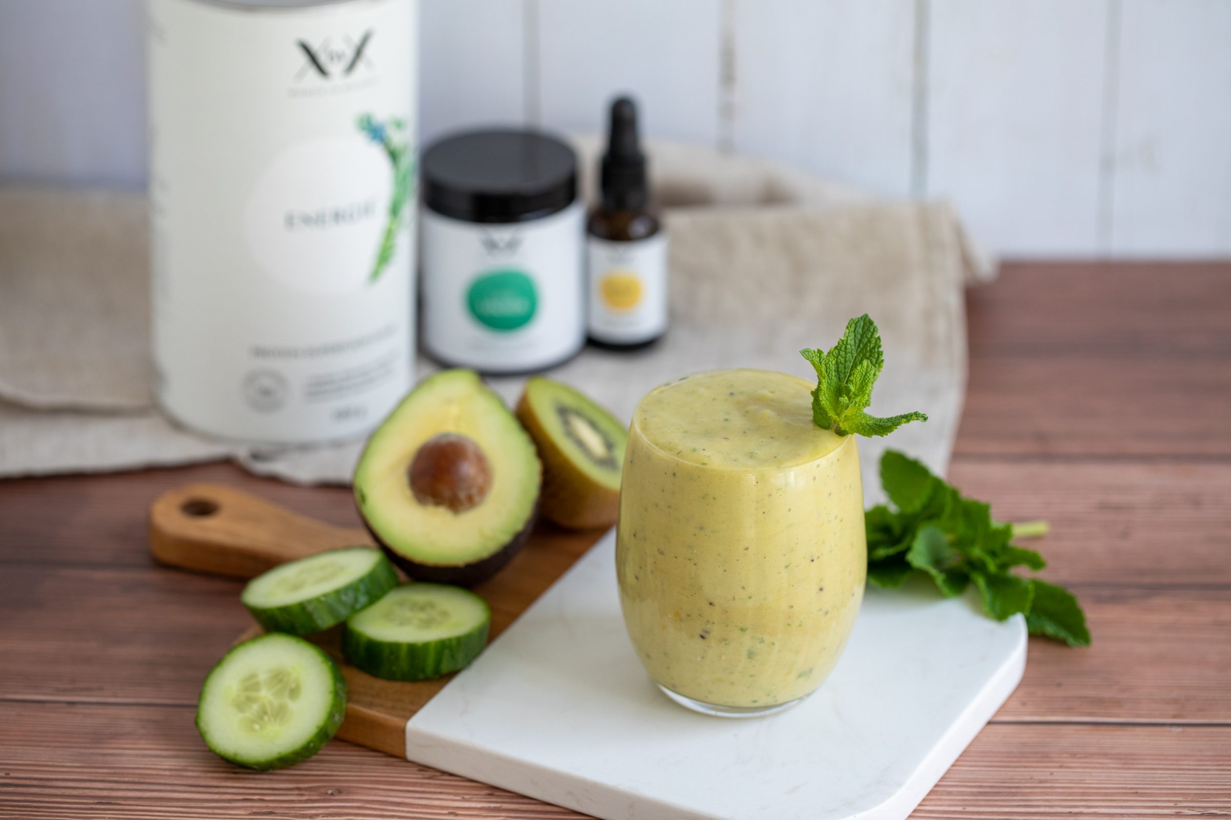 Grüner Smoothie im Glas neben Avocado, Gurke und Produktdosen von XbyX